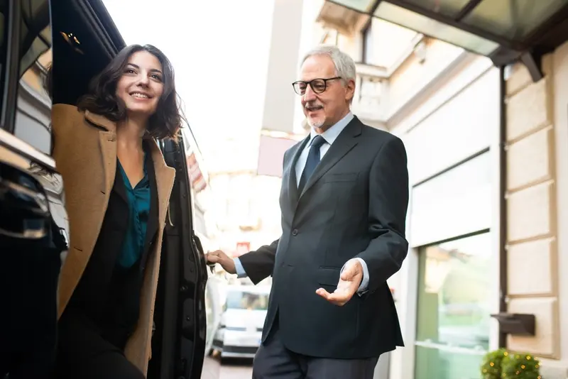 A professional chauffeur in a formal uniform opens the back door of a luxury black car for a stylish, smiling woman arriving at her destination, symbolizing premium Houston to Galveston transportation.